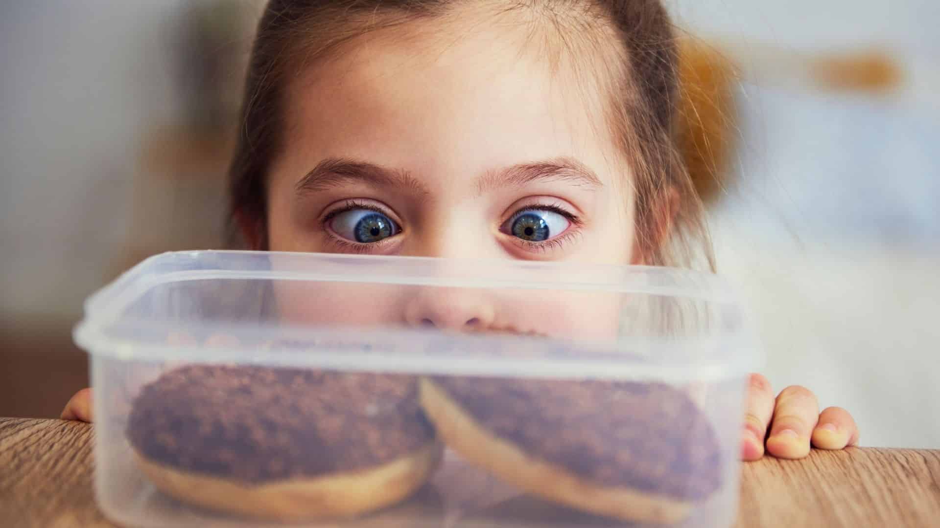 Kleines Mädchen schaut gierig auf 2 Donuts, die in einer Schale vor ihr liegen.