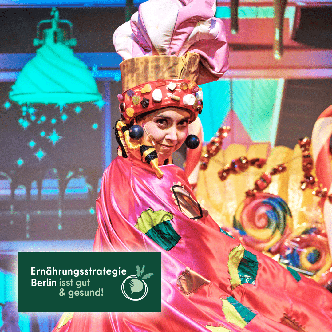 Eine Frau in einem pink-bunten Outfit mit Süßigkeiten am Kopfputz und Muffin-Motiven auf dem rosafarbenen Umhang. Im Hintergrund bunte Lutscher und Bonbons.