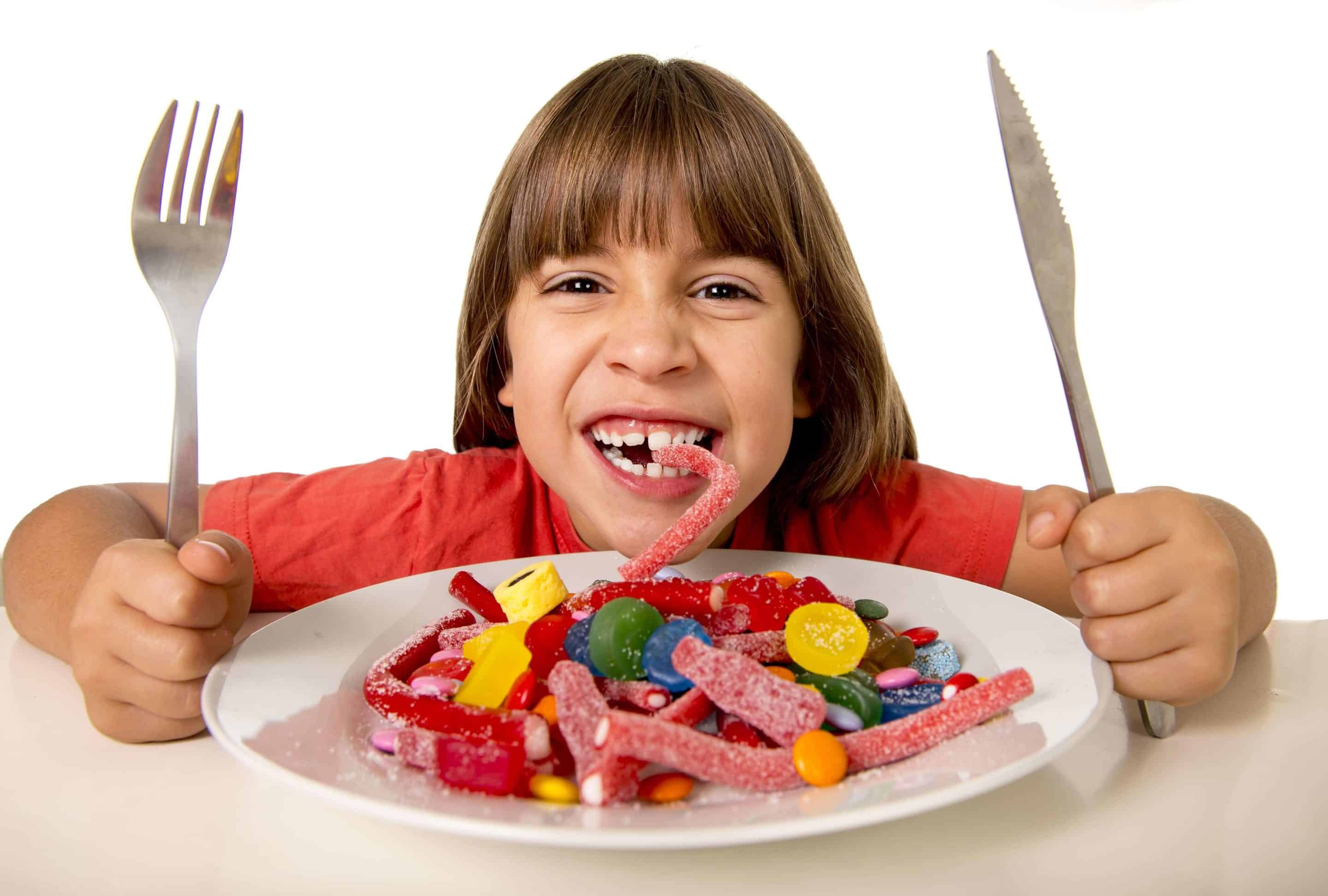Mädchen isst Fruchtgummis von einem mit Fruchtgummis gefülltem Teller und hält Messer und Gabel nach oben.