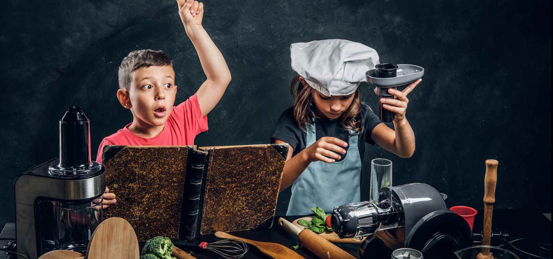 Zwei Kinder vor Utensilien aus der Küche. Ein Junge mit einem Kochbuch in der Hand, hat eine Idee. Ein Mädchen, mit einer Kochmütze auf dem Kopf, bedient eine Saftpresse.