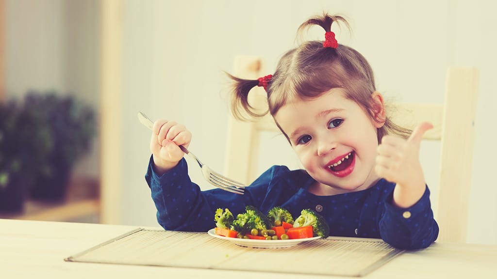 Kleines Mädchen mit 2 Zöpfen sitzt begeistert vor einem Teller mit Gemüse und zeigt einen Daumen hoch!